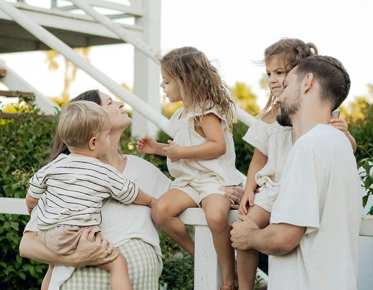 Parents holding their three young children by a white fence, the kids smiling up at them